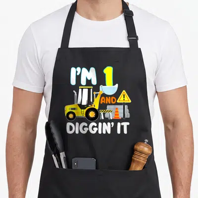 1st birthday truck apron - Front loader illustration with text I'M 1 AND DIGGIN IT and construction equipment in yellow — shown on a black cotton canvas apron against a white background