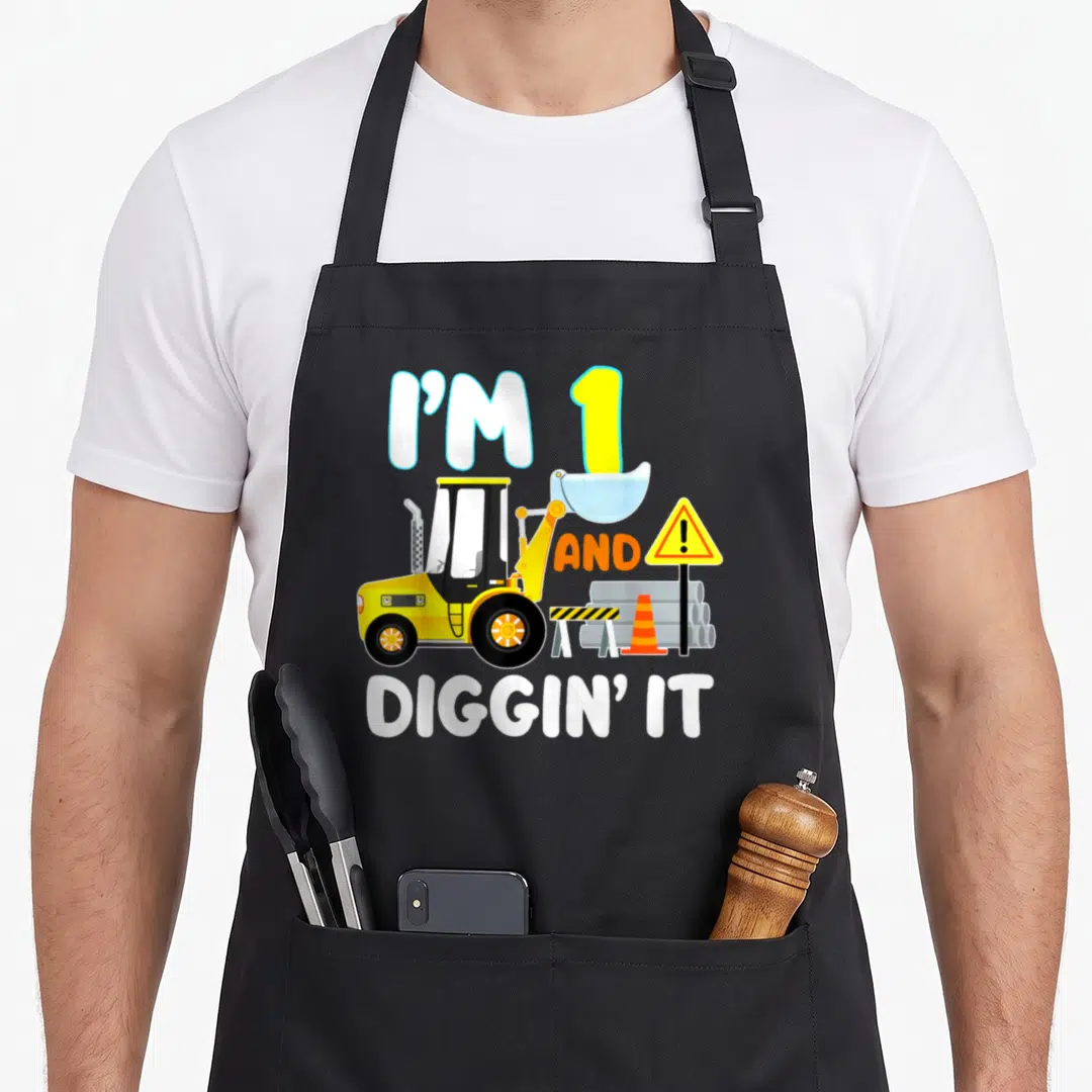 1st birthday truck apron - Front loader illustration with text I'M 1 AND DIGGIN IT and construction equipment in yellow — shown on a black cotton canvas apron against a white background