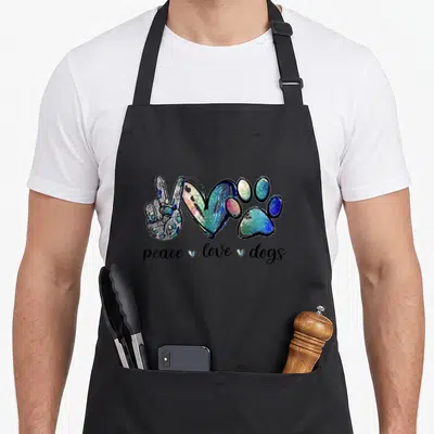 Peace dog apron - Peace sign hand, heart, and paw print with text peace love dogs in blue green pink and black — shown on a black cotton canvas apron against a white background