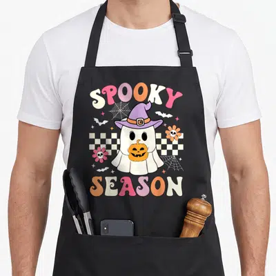 Groovy halloween apron - Retro cartoon ghost in purple hat holding pumpkin with SPOOKY SEASON text in pink, orange — shown on a black cotton canvas apron against a white background