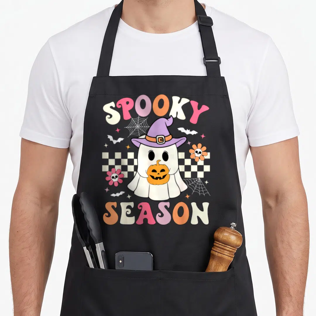 Groovy halloween apron - Retro cartoon ghost in purple hat holding pumpkin with SPOOKY SEASON text in pink, orange — shown on a black cotton canvas apron against a white background