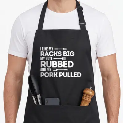 Rubbed barbecue apron - White stacked text reading I like my racks big my butt rubbed and my pork pulled with tools — shown on a black cotton canvas apron against a white background