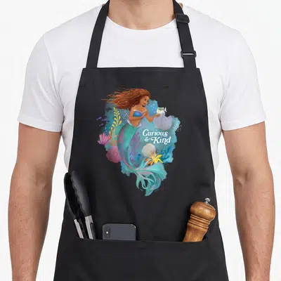 Curious mermaid apron - Watercolor mermaid with Curious & Kind text, teal blue pink orange coral sea life — shown on a black cotton canvas apron against a white background