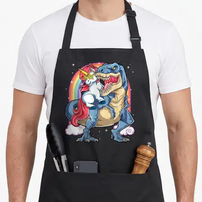 Riding rex rainbow dinosaur apron - Blue T-Rex holding white unicorn with red mane under rainbow arch with stars — shown on a black cotton canvas apron against a white background