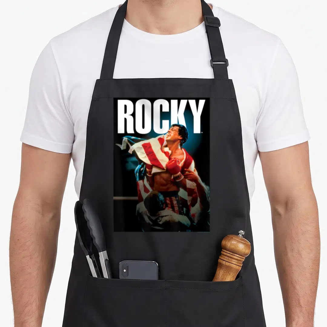 Rocky american flag apron - ROCKY text above boxer wrapped in flag with red glove — shown on a black cotton canvas apron against a white background