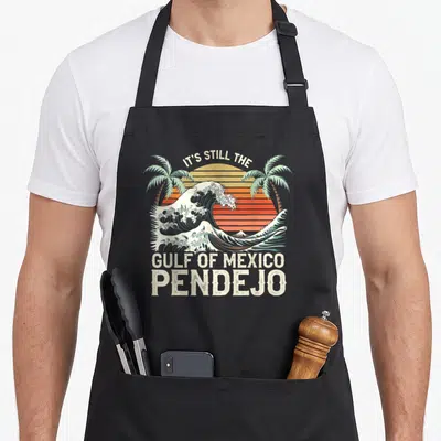Gulf spanish aprons - Retro wave and palm trees with text IT'S STILL THE GULF OF MEXICO PENDEJO — shown on a black cotton canvas apron against a white background