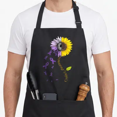 Disease sunflower apron - Split sunflower with purple ribbons and vertical Crohn's Awareness text — shown on a black cotton canvas apron against a white background