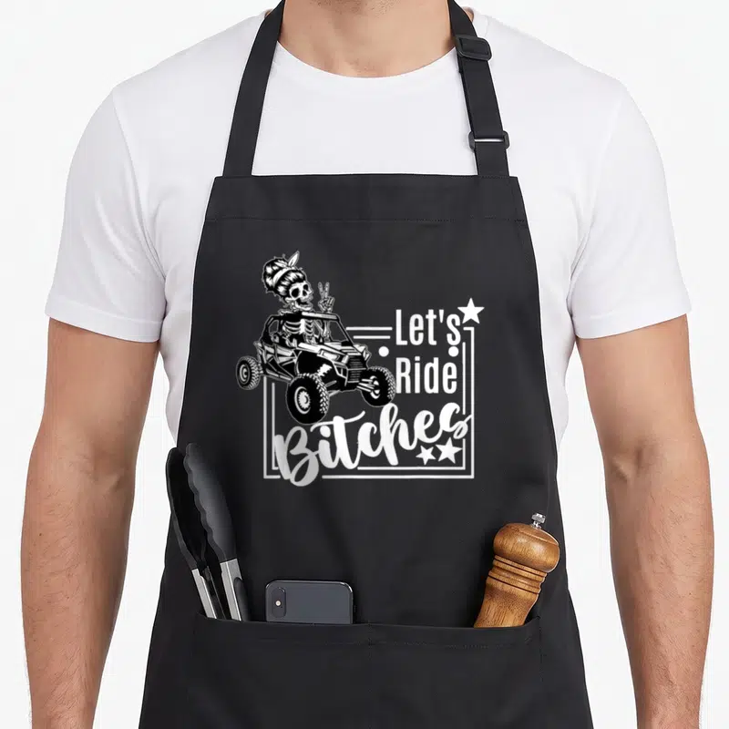 Ride utv girl apron - Skeleton in hair bun driving UTV with Let's Ride Bitches text and stars — shown on a black cotton canvas apron against a white background