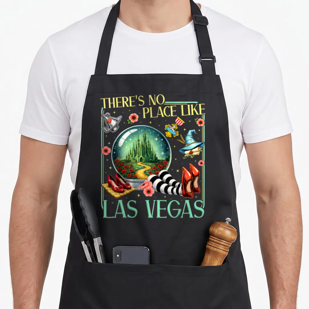 Brick wicked apron - Snow globe with green castle and yellow path surrounded by red shoes, witch hat, and text There's — shown on a black cotton canvas apron against a white background