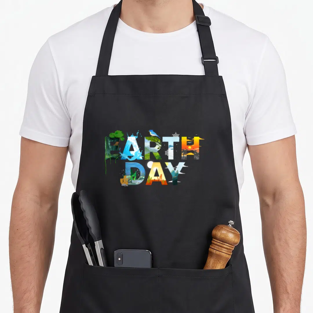 Environmental tree apron - Stacked EARTH DAY text filled with nature scenes in blue, green, yellow, and orange — shown on a black cotton canvas apron against a white background