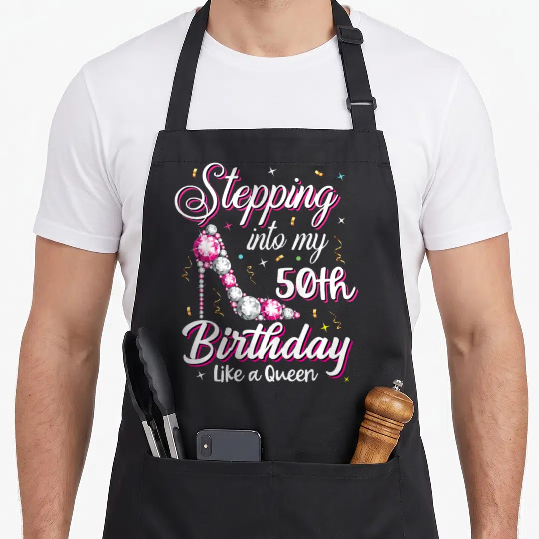 50th queen birthday apron - Jeweled high heel shoe with Stepping into my 50th Birthday Like a Queen text and confetti — shown on a black cotton canvas apron against a white background