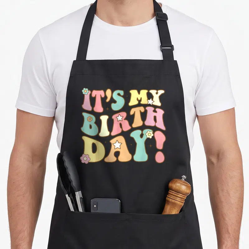 Toddler birthday apron - Retro IT'S MY BIRTH DAY! text with pink, yellow, blue, and orange flowers and stars — shown on a black cotton canvas apron against a white background