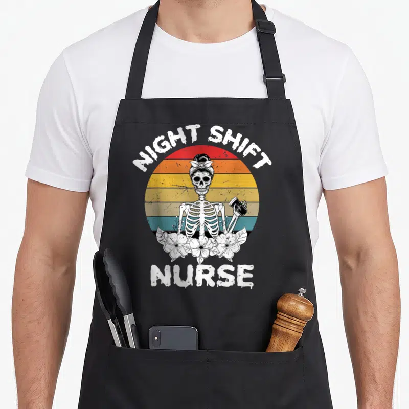 Night skeleton apron - Retro skeleton holding coffee cup with Night Shift Nurse text and striped sun background — shown on a black cotton canvas apron against a white background