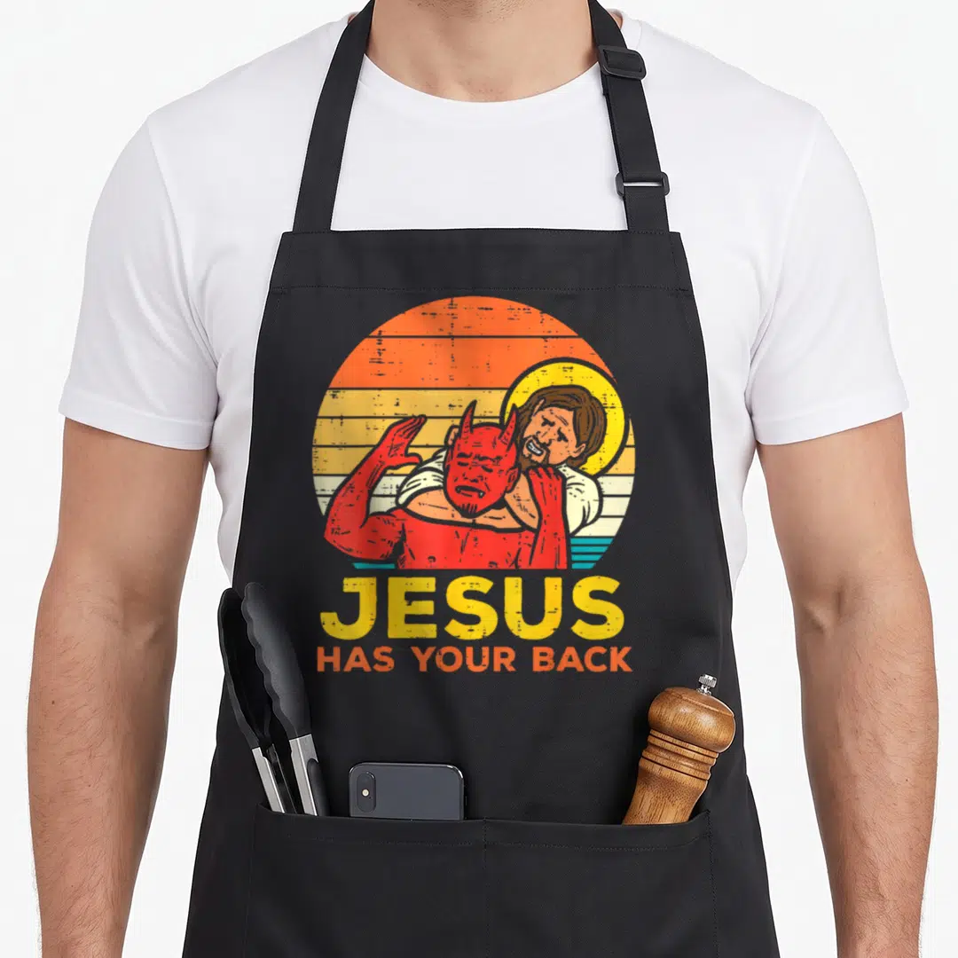 Jiu christian apron - Jesus holding red devil in headlock with stacked JESUS HAS YOUR BACK text on retro sun — shown on a black cotton canvas apron against a white background