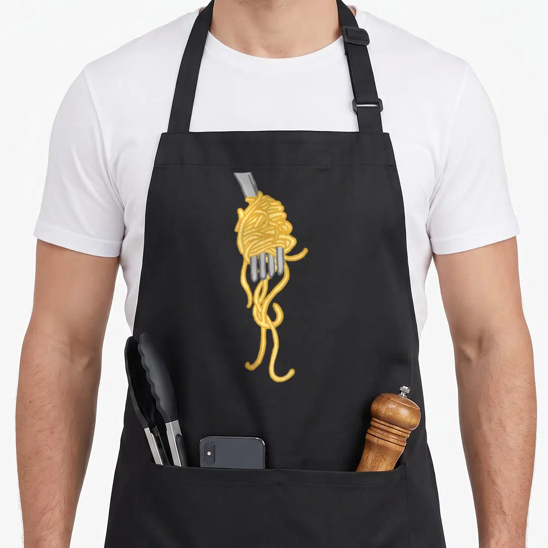 Gourmet pasta apron - Cartoon yellow spaghetti wrapped around silver fork tines with hanging strands — shown on a black cotton canvas apron against a white background