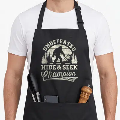 Undefeated hide bigfoot apron - Distressed off-white print of bigfoot silhouette with UNDEFEATED HIDE & SEEK Champion — shown on a black cotton canvas apron against a white background