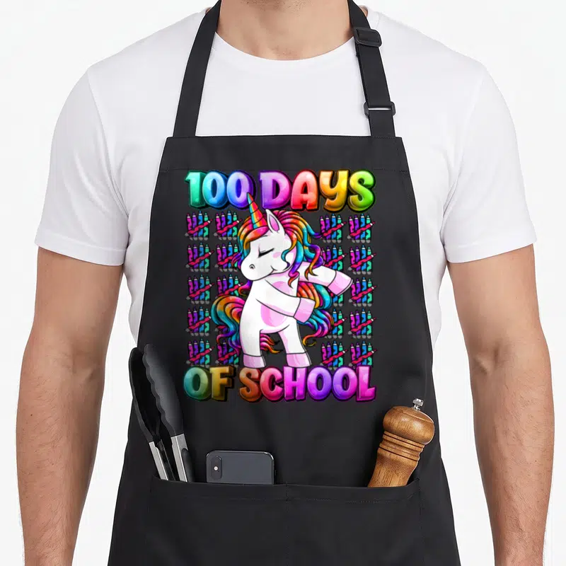 School smarter unicorn apron - Cartoon unicorn dabbing on grid of crayons with 100 Days of School text — shown on a black cotton canvas apron against a white background
