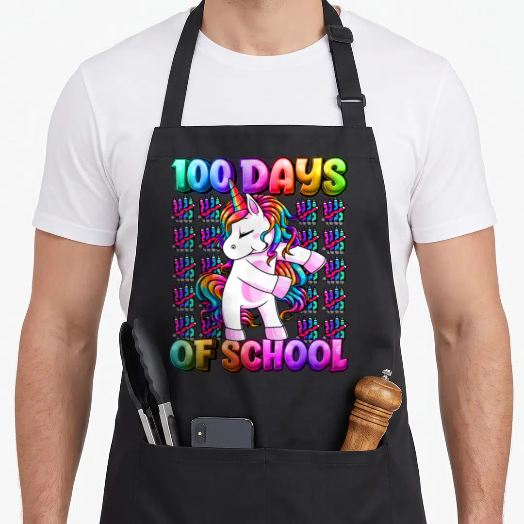 School smarter unicorn apron - Cartoon unicorn dabbing on grid of crayons with 100 Days of School text — shown on a black cotton canvas apron against a white background