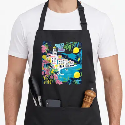 Bella italy apron - Cartoon coastal village with lemons pink flowers and Ciao Bella text in blue bubble — shown on a black cotton canvas apron against a white background