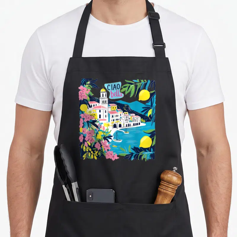 Bella italy apron - Cartoon coastal village with lemons pink flowers and Ciao Bella text in blue bubble — shown on a black cotton canvas apron against a white background