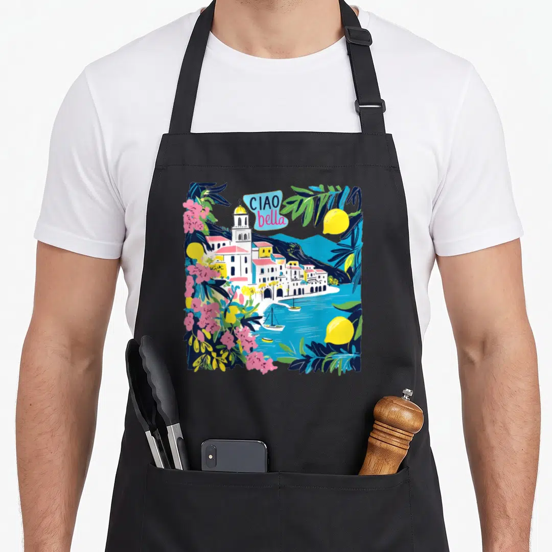 Bella italy apron - Cartoon coastal village with lemons pink flowers and Ciao Bella text in blue bubble — shown on a black cotton canvas apron against a white background