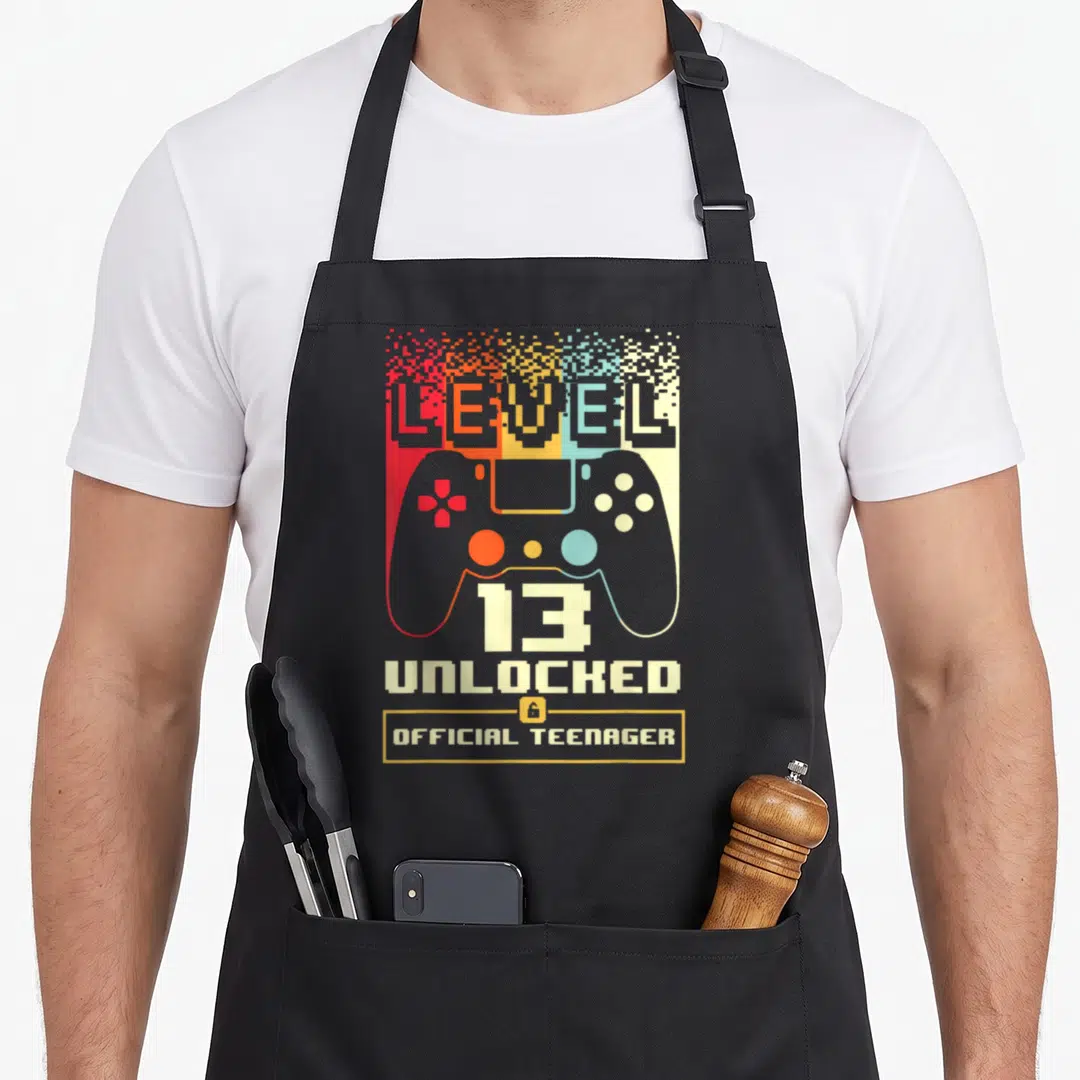 13th level birthday apron - Pixelated rainbow game controller with text Level 13 Unlocked Official Teenager — shown on a black cotton canvas apron against a white background