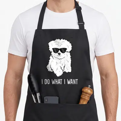 Maltese owner dog apron - Cartoon white Maltese dog wearing black sunglasses above text I DO WHAT I WANT — shown on a black cotton canvas apron against a white background