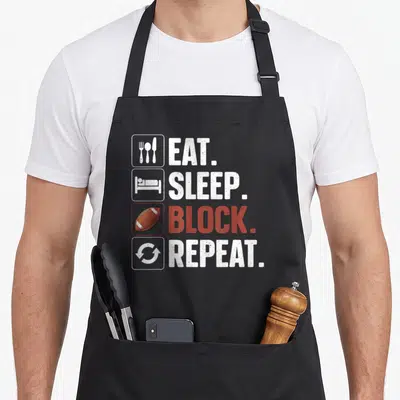 Lineman player football apron - EAT SLEEP BLOCK REPEAT text with cutlery bed football and repeat icons in white and red — shown on a black cotton canvas apron against a white background