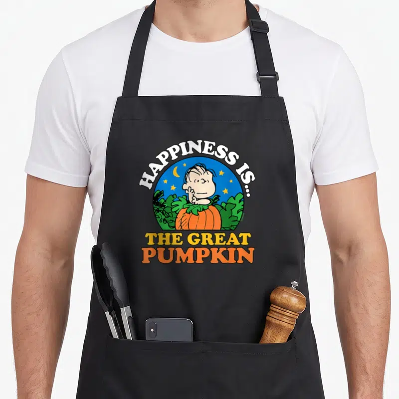 Linus pumpkin apron - Linus behind orange pumpkin in blue circle with stars and arched white text reading Happiness Is — shown on a black cotton canvas apron against a white background