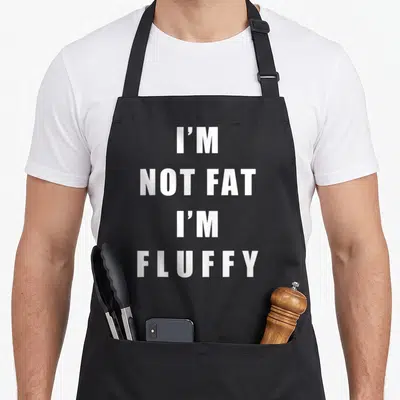 Fluffy fat man in apron - White stacked text I'M NOT FAT I'M FLUFFY on black bib apron — shown on a black cotton canvas apron against a white background