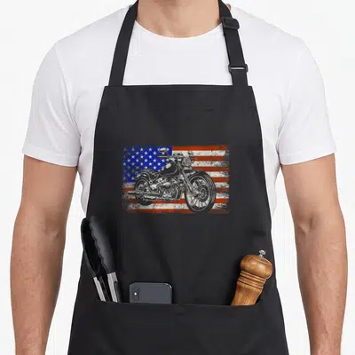 Riding american flag apron - White motorcycle silhouette on distressed red white and blue American flag background — shown on a black cotton canvas apron against a white background