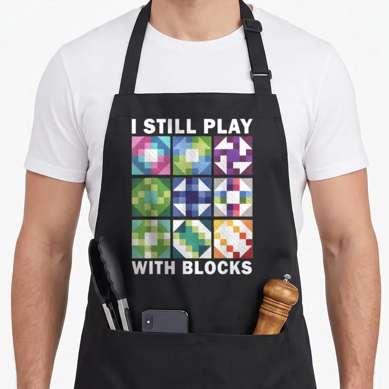 Quilting sewing quilter apron - I STILL PLAY WITH BLOCKS text above 3x3 grid of colorful geometric quilting squares — shown on a black cotton canvas apron against a white background