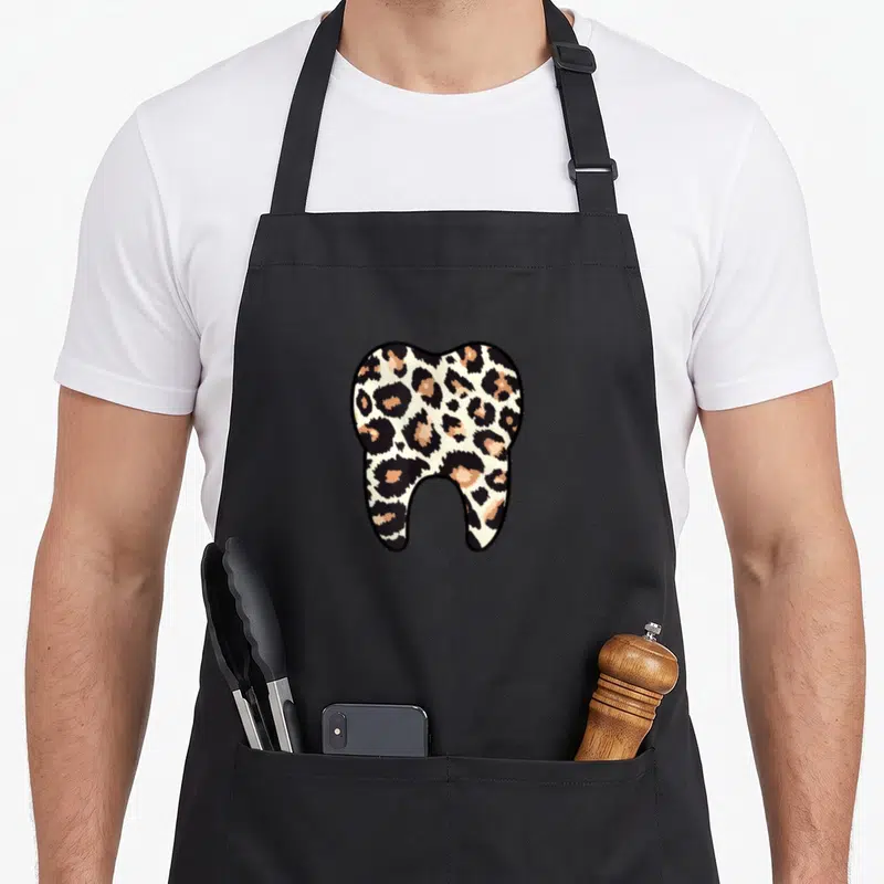 Leopard dentist apron - Cartoon molar filled with tan black and white leopard spots — shown on a black cotton canvas apron against a white background