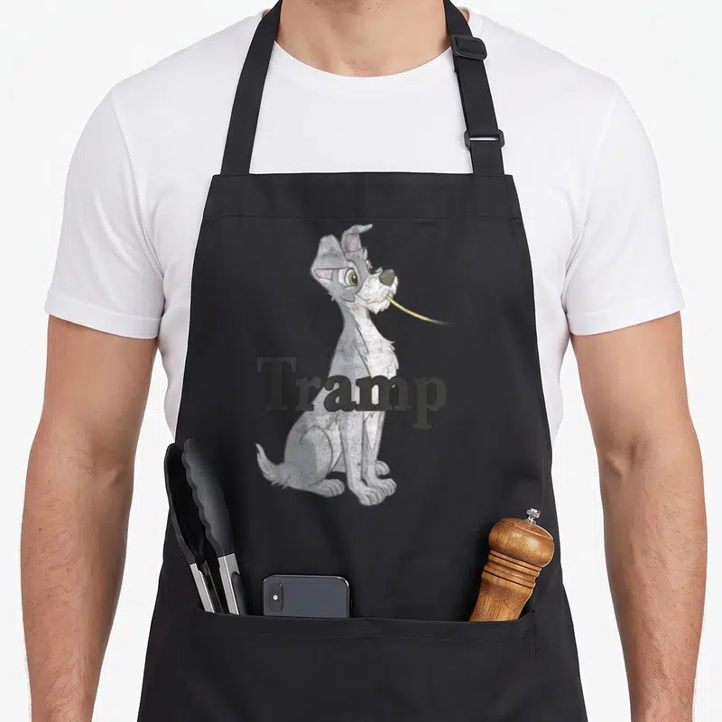 Couple lady and the tramp apron - Grey cartoon dog holding yellow noodle with Tramp text overlay — shown on a black cotton canvas apron against a white background