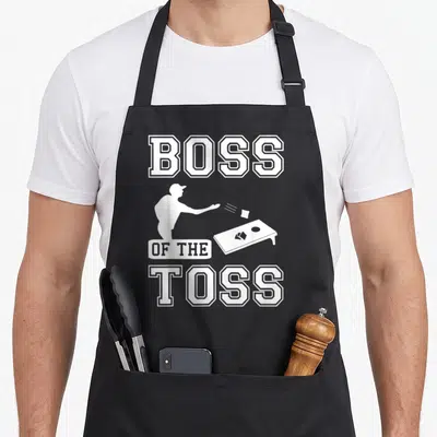 Toss boss apron - White Boss of the Toss text and cornhole player silhouette on grey background — shown on a black cotton canvas apron against a white background