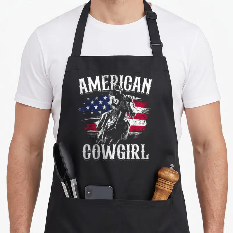 Rodeo cowgirl aprons - Arched American Cowgirl text framing a distressed US flag and horse rider silhouette — shown on a black cotton canvas apron against a white background