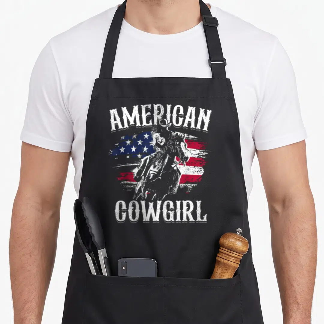 Rodeo cowgirl aprons - Arched American Cowgirl text framing a distressed US flag and horse rider silhouette — shown on a black cotton canvas apron against a white background