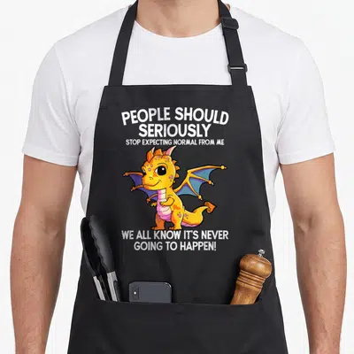 Dragon apron for men - Cartoon yellow dragon surrounded by white text about not expecting normal — shown on a black cotton canvas apron against a white background