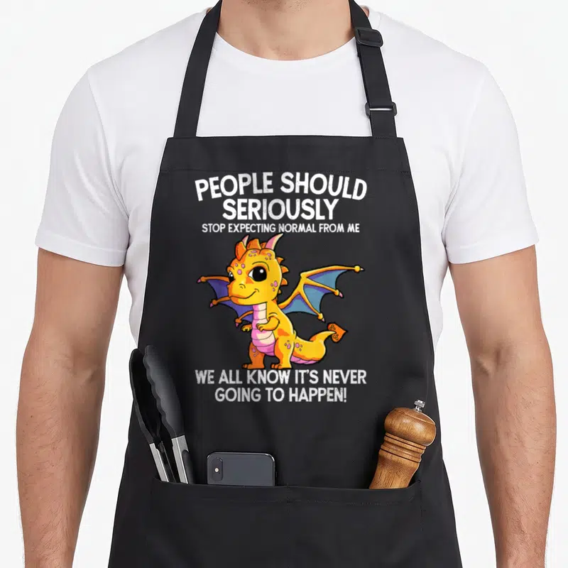 Dragon apron for men - Cartoon yellow dragon surrounded by white text about not expecting normal — shown on a black cotton canvas apron against a white background