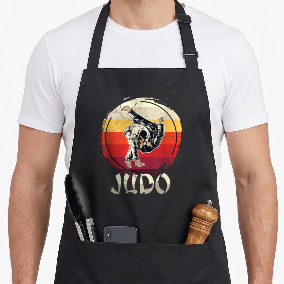 Judo modern apron - Retro sunset circle with judo throw and JUDO lettering in red orange beige — shown on a black cotton canvas apron against a white background