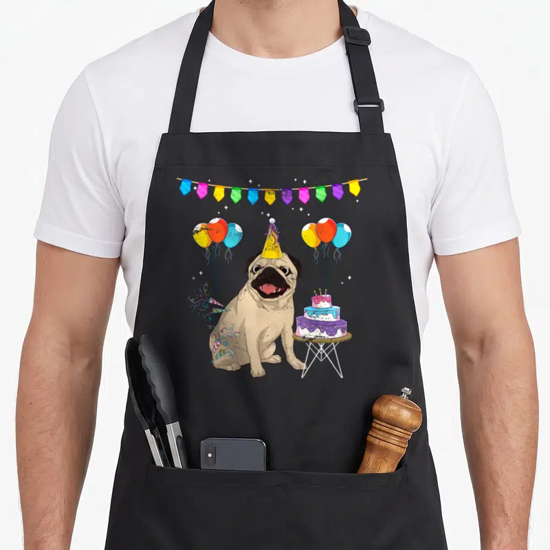 Cake dog apron - Cartoon pug in party hat beside tiered cake with balloons and bunting in tan, yellow, blue, pink — shown on a black cotton canvas apron against a white background