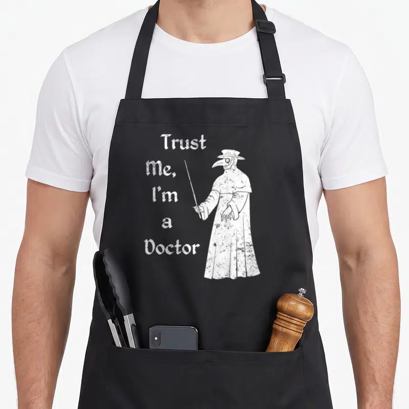 Trust plague doctor apron - White distressed text Trust Me I'm a Doctor beside a plague doctor figure pointing a cane — shown on a black cotton canvas apron against a white background