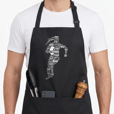Proud rugby apron - White rugby player silhouette formed by words like Haka, Tackle, Flyhalf, and Press — shown on a black cotton canvas apron against a white background