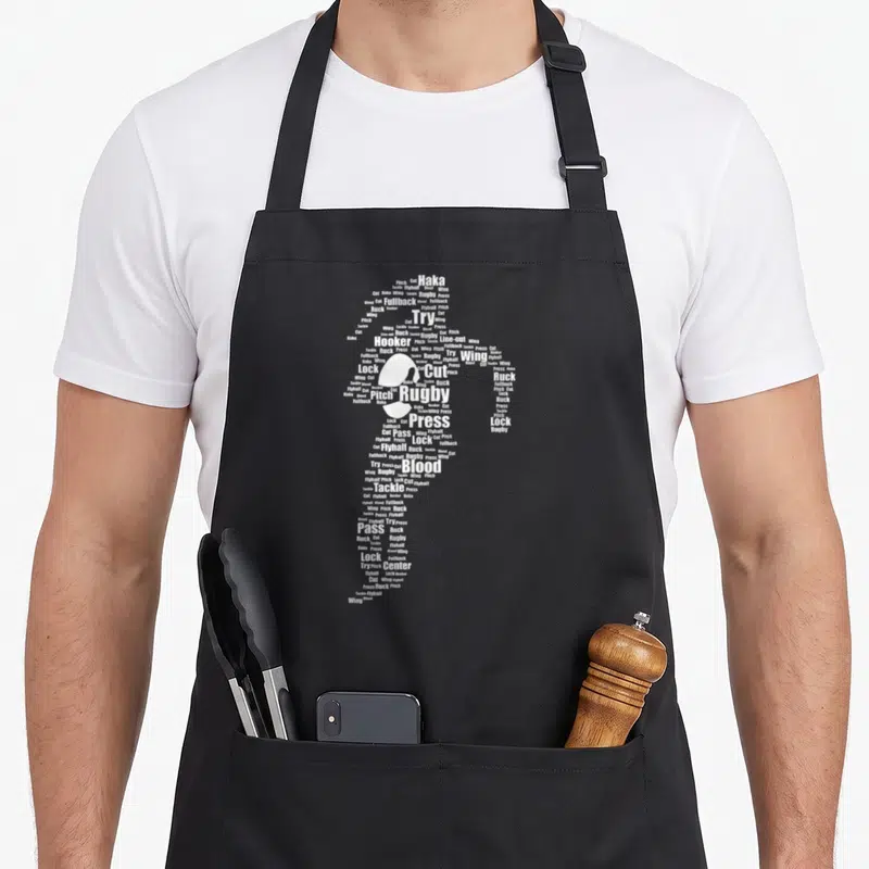 Proud rugby apron - White rugby player silhouette formed by words like Haka, Tackle, Flyhalf, and Press — shown on a black cotton canvas apron against a white background