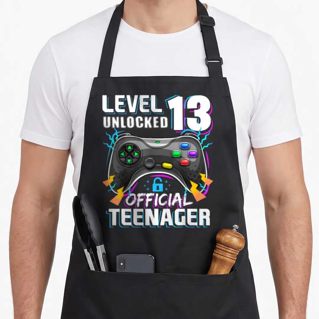 13th video game apron - Level 13 Unlocked Official Teenager text with controller, blue lock, and lightning bolts — shown on a black cotton canvas apron against a white background