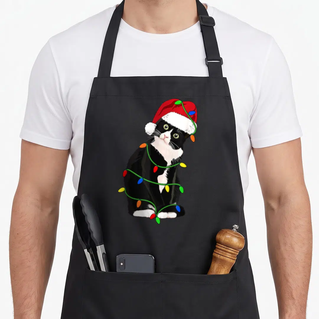 Meow tuxedo apron - Cartoon tuxedo cat in red santa hat wrapped in green string lights with multicolored bulbs — shown on a black cotton canvas apron against a white background