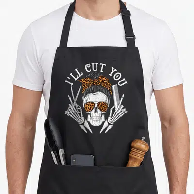 Ill skull apron - Cartoon skull with leopard print bow and sunglasses flanked by skeleton hands holding scissors — shown on a black cotton canvas apron against a white background