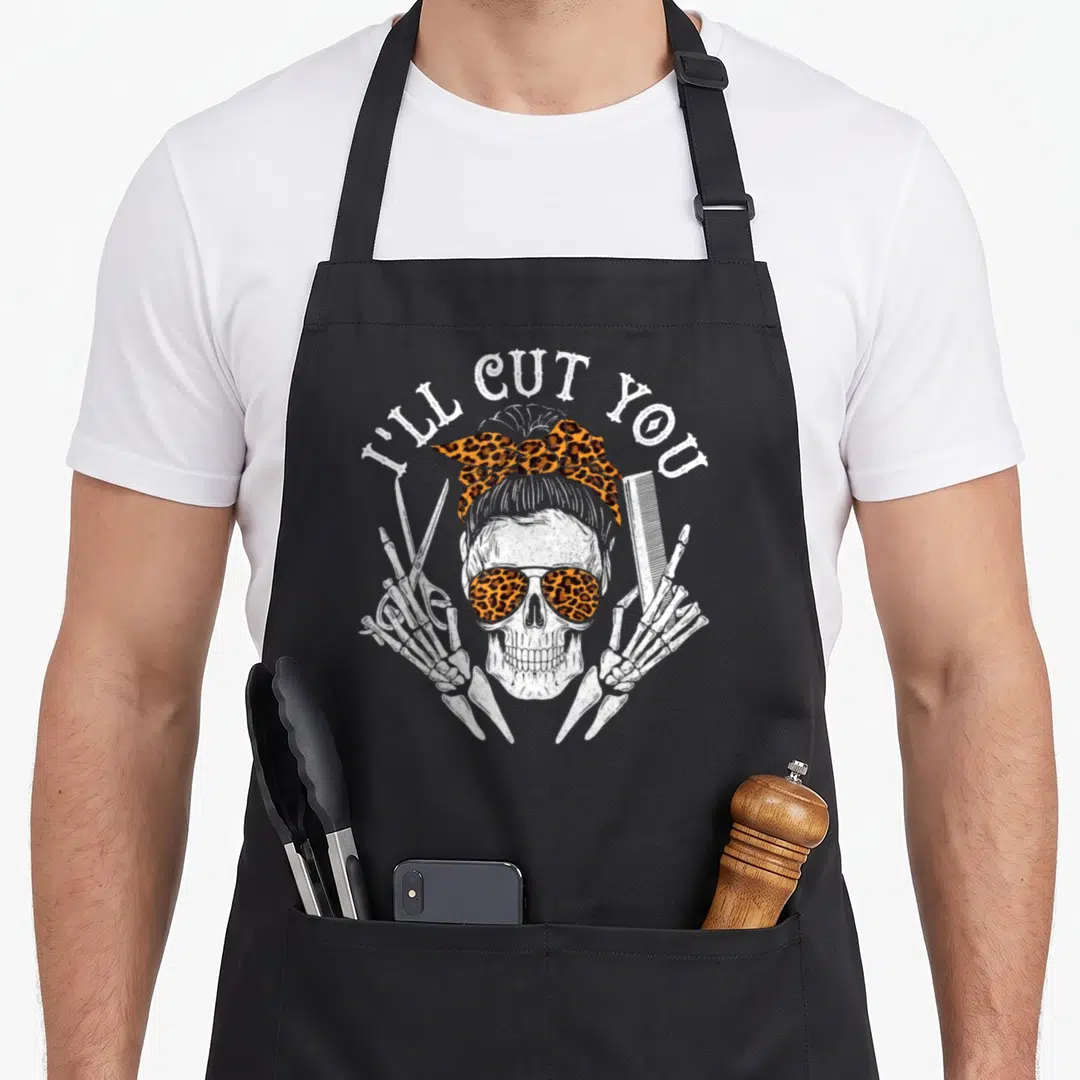 Ill skull apron - Cartoon skull with leopard print bow and sunglasses flanked by skeleton hands holding scissors — shown on a black cotton canvas apron against a white background
