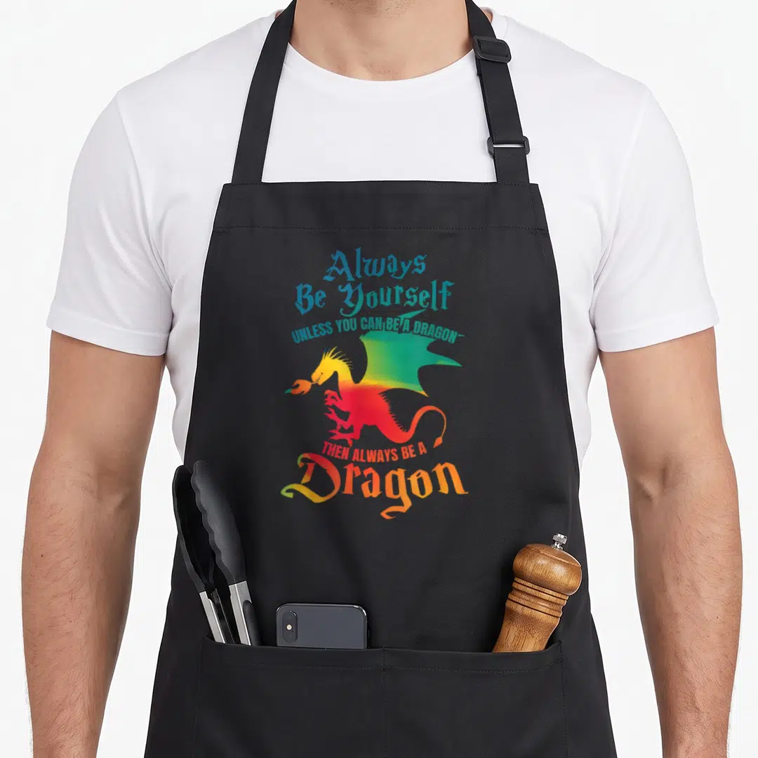 Alway dragon apron - Cartoon dragon in red yellow green blue with gothic text Always Be Yourself Unless You Can Be — shown on a black cotton canvas apron against a white background