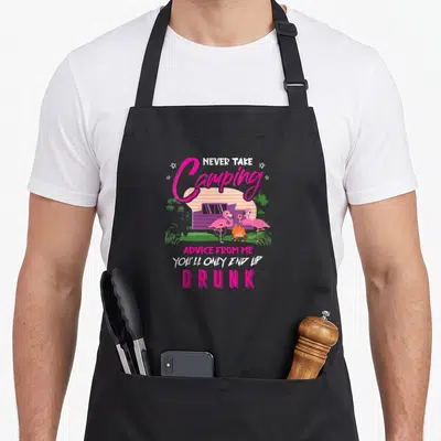 Youll camping apron - Two pink flamingos holding wine glasses by a campfire in front of a purple camper van with text — shown on a black cotton canvas apron against a white background